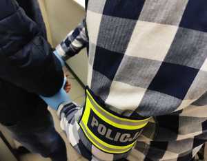 obraz przedstawia ramię policjanta z opaską z napisem policja zapinającego kajdanki na dłonie zatrzymanego