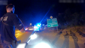 Policjant idzie poboczem i świeci latarką. W tle widać zaparkowany radiowóz policyjny z włączonymi światłami błyskowymi.