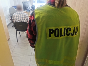 Zatrzymany mężczyzna siedzi na krześle, za nim stoi policjantka w cywilnym ubraniu, w kamizelce z napisem policja.